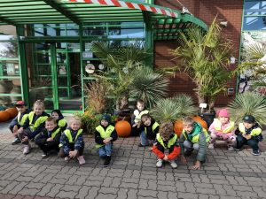 Przedszkolaki pozują do wspólnego zdjęcia przed wejściem do centrum ogrodniczego, wśród palm, roślin ozdobnych i dużych pomarańczowych dyń.