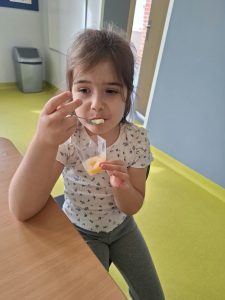 Dziewczynka siedząca przy stole próbuje marmolady z przezroczystego kubeczka. Trzyma łyżeczkę przy ustach i skupia się na degustacji przygotowanego przysmaku.
