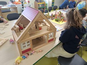 Drewniany domek dla lalek z mebelkami stoi na stoliku w sali przedszkolnej. W tle dzieci bawią się przy innych stanowiskach.
