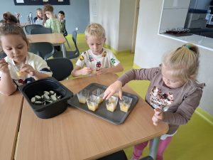 Troje przedszkolaków siedzi przy stole i degustuje domową marmoladę z małych przezroczystych kubeczków. Na tacy stoją kolejne porcje przysmaku, a obok znajduje się pojemnik z łyżeczkami.