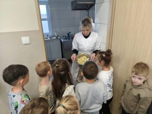 Pani w białym fartuchu pokazuje przedszkolakom garnek wypełniony obranymi i pokrojonymi na kawałki owocami. Dzieci stoją przed wejściem do szkolnej kuchni i przyglądają się przygotowanym składnikom marmolady.