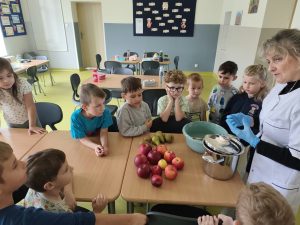 Dzieci z oddziału przedszkolnego zgromadzone wokół stołu obserwują przygotowania do wykonania domowej marmolady. Pani w białym fartuchu pokazuje produkty i przybory kuchenne. Na stole leżą jabłka, gruszki, miska oraz duży garnek.