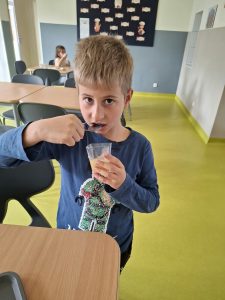 Chłopiec w niebieskiej bluzce próbuje przygotowanej marmolady, nabierając ją łyżeczką z przezroczystego kubeczka. W tle widoczna jest szkolna jadalnia.