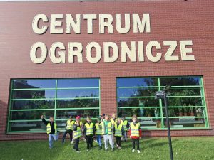Grupa przedszkolaków w kamizelkach odblaskowych pozuje przed budynkiem Centrum Ogrodniczego w Ostaszewie, na którego elewacji widnieje duży napis „CENTRUM OGRODNICZE”.