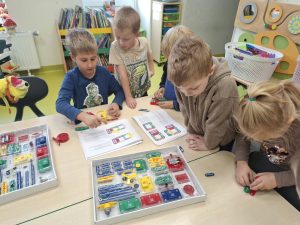 Grupa przedszkolaków przy stoliku wspólnie poznaje elementy zestawu do budowania obwodów elektrycznych, korzystając z instrukcji obrazkowej.