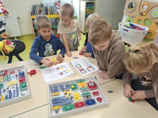 Grupa przedszkolaków przy stoliku wspólnie poznaje elementy zestawu do budowania obwodów elektrycznych, korzystając z instrukcji obrazkowej.