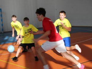 Uczniowie w czerwonych i żółtych strojach podczas dynamicznej akcji w meczu piłki ręcznej.
