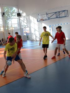 Uczniowie w czerwonych i żółtych strojach walczą o piłkę podczas meczu piłki ręcznej.