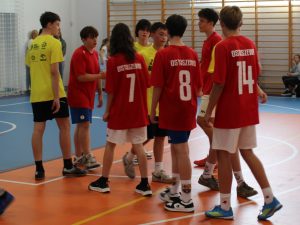 Uczniowie w czerwonych i żółtych strojach sportowych zgromadzeni na boisku podczas meczu piłki ręcznej.