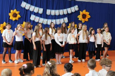 Uczennice szkolnego chórku podczas występu z okazji Dnia Edukacji Narodowej. W tle granatowa dekoracja z napisem „Dzień Edukacji Narodowej” i żółtymi słonecznikami.