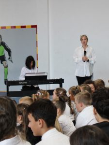 Uczeń gra na pianinie cyfrowym, a stojąca obok nauczycielka trzyma mikrofon. Na pierwszym planie siedzą uczniowie uczestniczący w akademii.