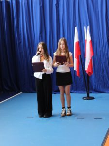 Dwie uczennice prowadzą uroczystą akademię. Jedna przemawia do mikrofonu, a obie trzymają bordowe teczki. Obok stoją polskie flagi.