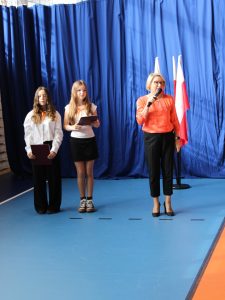 Pani dyrektor w pomarańczowej bluzce przemawia do mikrofonu. Obok stoją dwie uczennice prowadzące akademię, trzymające teczki.