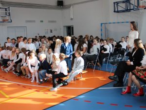 Uczniowie różnych klas zgromadzeni w sali gimnastycznej podczas uroczystości z okazji Dnia Edukacji Narodowej. Po prawej stronie siedzą nauczyciele. Uczniowie klasy II i ich wychowawczynia stoją.