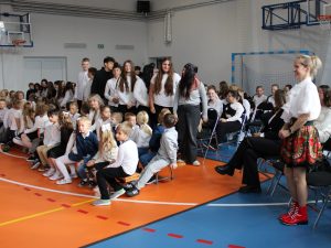 Grupa starszych uczniów stoi wśród zgromadzonych uczestników akademii. Na pierwszym planie siedzą młodsze dzieci, a po prawej nauczycielki. Uczniowie klasy VII i ich wychowawczynia stoją.