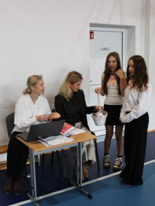 Dwie uczennice z białym koszyczkiem wręczają słodkie upominki nauczycielkom siedzącym przy stoliku z laptopem i dokumentami.