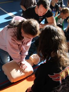 Strażak w czarnej koszulce obserwuje uczennice ćwiczące resuscytację na fantomie i udziela im wskazówek.