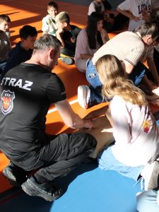 Strażak z Ochotniczej Straży Pożarnej pomaga uczennicy prawidłowo ułożyć dłonie podczas ćwiczenia ucisków klatki piersiowej na fantomie.