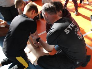 Uczniowie ćwiczą resuscytację na fantomach pod opieką dorosłych. Strażak pomaga chłopcu przygotować się do wykonania ucisków klatki piersiowej.