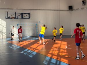 Uczniowie w czerwonych i żółtych strojach podczas meczu piłki ręcznej; zawodnik w żółtej koszulce wyskakuje do podania piłki.