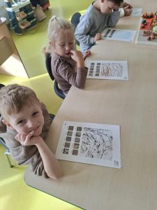 Przedszkolaki siedzą przy stoliku i oglądają wykonane przez siebie odbitki kory drzew. Na kartach widoczne są różnokolorowe wzory odzwierciedlające fakturę pni.