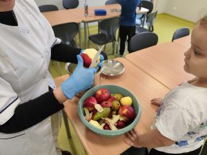 Pani w białym fartuchu i niebieskich rękawiczkach obiera jabłko nożem. Obok stoi dziewczynka, która uważnie obserwuje pracę. Na stole znajduje się miska z jabłkami, gruszkami i obierkami.