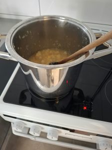 Metalowy garnek stojący na kuchence elektrycznej, wypełniony gotującą się masą owocową. W garnku znajduje się drewniana łyżka służąca do mieszania przygotowywanej marmolady.