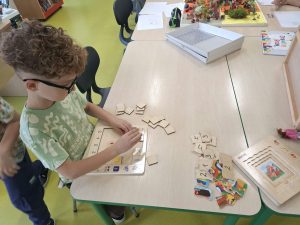 Chłopiec w okularach dopasowuje drewniane elementy do planszy edukacyjnej. Na stoliku leżą również inne układanki i puzzle.