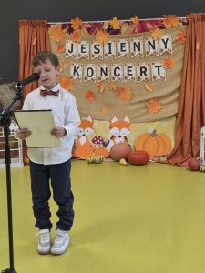 Chłopiec w białej koszuli i brązowej muszce czyta tekst z kartki do mikrofonu na tle dekoracji z napisem „Jesienny koncert”.