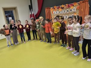 Grupa dzieci w jesiennych strojach i przebraniach zwierząt śpiewa i wykonuje gesty podczas wspólnego występu.