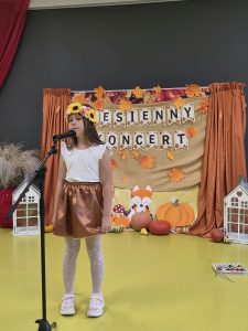 Dziewczynka w białej bluzce, brązowej spódniczce i wianku ze słoneczników śpiewa do mikrofonu przed dekoracją „Jesienny koncert”.