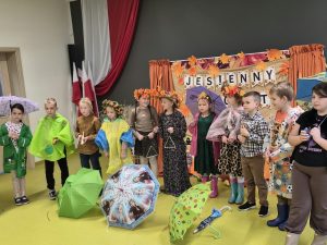 Grupa dzieci w jesiennych przebraniach i płaszczach przeciwdeszczowych stoi na scenie wśród kolorowych parasoli. Niektóre dzieci trzymają instrumenty muzyczne.