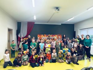 Uczniowie klas 0–III wraz z nauczycielami pozują do wspólnego zdjęcia po jesiennym koncercie. Dzieci mają na sobie kolorowe stroje i przebrania związane z jesienią.