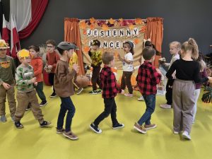 Dzieci w jesiennych przebraniach tańczą i poruszają się po scenie podczas wspólnego występu na tle dekoracji „Jesienny koncert”.