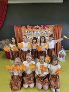 Osiem dziewczynek w białych bluzkach i brązowych spódniczkach pozuje z pomarańczowymi liśćmi na tle dekoracji „Jesienny koncert”.