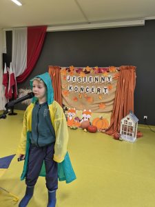 Chłopiec w kolorowym płaszczu przeciwdeszczowym i kaloszach występuje przy mikrofonie na tle jesiennej scenografii.