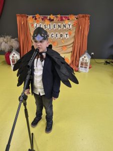 Chłopiec przebrany za czarnego ptaka, z maską na głowie i skrzydłami, recytuje do mikrofonu podczas jesiennego koncertu.