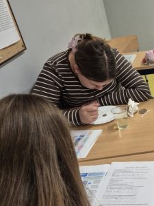 Uczennica klasy VII pochyla się nad monetą umieszczoną na talerzyku i za pomocą pipety nanosi na nią krople wody. Na ławce leżą karta pracy i materiały do doświadczenia.