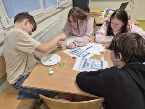 Grupa uczniów klasy VII pracuje przy wspólnym stole podczas lekcji fizyki. Jeden z chłopców miesza ciecz w przezroczystym kubeczku, a pozostali obserwują przygotowania do doświadczenia.