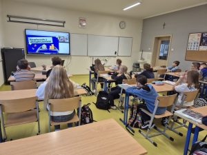 Uczniowie klasy szkolnej uczestniczą w lekcji online poświęconej finansom. Na monitorze interaktywnym widoczna jest prezentacja zatytułowana „Bezpiecznie z ubezpieczeniami”.
