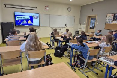 Uczniowie klasy szkolnej uczestniczą w lekcji online poświęconej finansom. Na monitorze interaktywnym widoczna jest prezentacja zatytułowana „Bezpiecznie z ubezpieczeniami”.