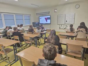 Uczniowie siedzą w szkolnych ławkach i oglądają prezentację podczas lekcji online poświęconej funkcjonowaniu spółdzielni uczniowskiej.