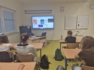 Uczniowie uczestniczą w lekcji online dotyczącej edukacji finansowej. Na monitorze interaktywnym widoczne są materiały edukacyjne, w tym dotyczące matematyki i geografii.