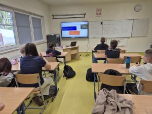 Uczniowie siedzą w ławkach i uczestniczą w lekcji online dotyczącej ekonomii. Na monitorze interaktywnym wyświetlana jest prezentacja prowadzącej spotkanie.