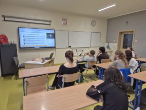 Uczniowie uczestniczą w lekcji online poświęconej inteligencji finansowej. Na monitorze widoczna jest lista zagadnień dotyczących budżetu, oszczędzania i przedsiębiorczości.