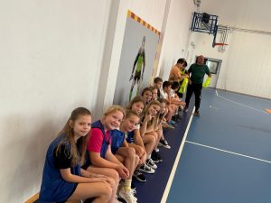 Uczniowie siedzą przy ścianie sali gimnastycznej i obserwują rozgrywane zawody sportowe.