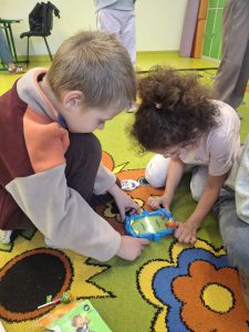 Dwoje dzieci z klasy I siedzi na kolorowym dywanie i wspólnie bawi się niewielką grą zręcznościową.