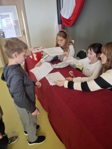 Uczeń klasy I odbiera kartę do głosowania od uczennic zasiadających przy stole komisji wyborczej.