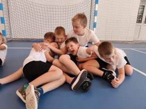 Grupa uśmiechniętych uczniów klasy I w białych koszulkach bawi się wspólnie na podłodze sali gimnastycznej.