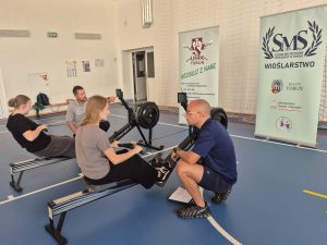 Dwie uczennice ćwiczą na ergometrach wioślarskich pod okiem trenerów; w tle widoczne są banery AZS UMK Toruń i Szkoły Mistrzostwa Sportowego w Toruniu.